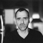 Imagem. Início da descrição de imagem: Retrato frontal em preto e branco de um homem de pele clara, com barba por fazer e cabelos curtos e escuros. Ele veste uma camiseta de gola em "V" escura e olha diretamente para a câmera com uma expressão séria e compenetrada. A fotografia possui uma granulação artística acentuada, e o fundo está bastante desfocado, sugerindo um ambiente interno doméstico com luzes suaves ao fundo. Fim da descrição de imagem.