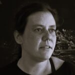 Imagem. Início da descrição de imagem: Retrato frontal em plano fechado (close-up) e tom sépia de uma mulher de pele clara, com cabelos lisos, escuros, curtos e levemente bagunçados. Ela olha para a direita da câmera com uma expressão serena e contemplativa. Sua pele apresenta marcas sutis como sardas. Veste uma camiseta de gola redonda simples em tom escuro. O fundo da imagem remete a detalhes de uma obra de arte com aspecto espinhoso. Fim da descrição de imagem.