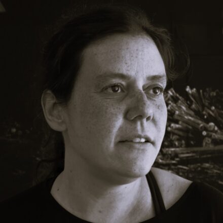 Imagem. Início da descrição de imagem: Retrato frontal em plano fechado (close-up) e tom sépia de uma mulher de pele clara, com cabelos lisos, escuros, curtos e levemente bagunçados. Ela olha para a direita da câmera com uma expressão serena e contemplativa. Sua pele apresenta marcas sutis como sardas. Veste uma camiseta de gola redonda simples em tom escuro. O fundo da imagem remete a detalhes de uma obra de arte com aspecto espinhoso. Fim da descrição de imagem.