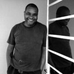 Imagem. Início da descrição de imagem: Retrato em preto e branco de um homem negro de pele retinta, com cabelos curtos e barba rala. Ele está posicionado em pé, ligeiramente à esquerda do centro, olhando diretamente para a câmera com um sorriso aberto e acolhedor. Veste uma camiseta escura de gola redonda que apresenta algumas manchas claras (possivelmente de tinta ou gesso), sugerindo um ambiente de trabalho criativo. Suas mãos estão nos bolsos. À direita da imagem, há uma superfície reflexiva com barras horizontais brancas, que projeta um reflexo da silhueta do artista, criando um jogo de duplicação e profundidade. O fundo à esquerda é liso e claro, mantendo o foco na expressão e na presença do retratado. Fim da descrição de imagem.