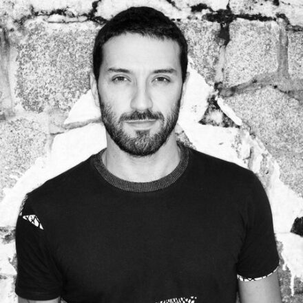 Imagem. Início da descrição de imagem: Retrato em preto e branco de um homem de pele clara com barba curta e cabelo escuro, olhando diretamente para a câmera com uma expressão séria. Ele veste uma camiseta preta com estampas geométricas em branco na área do peito e possui tatuagens visíveis no braço. O fundo é um muro de pedra de aparência antiga, com partes da argamassa descascada. Fim da descrição de imagem.