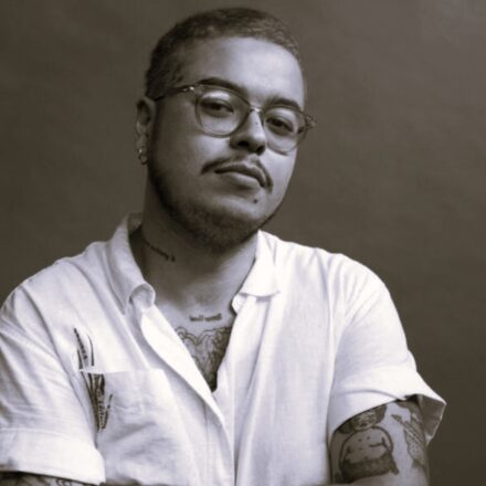 Descrição da imagem: a imagem é um retrato em preto e branco de um homem branco. O homem está em primeiro plano, olhando diretamente para a câmera, com uma expressão séria. Ele tem cabelo curto, barba e bigode finos, e usa óculos de aro claro. Ele veste uma camisa branca de botão com as mangas curtas e arregaçadas e está com os braços cruzados na frente do peito. Muitas tatuagens são visíveis, especialmente no braço esquerdo e no peito (incluindo uma que se estende para o pescoço). Ele também usa um brinco pequeno de argola em na orelha. O fundo é neutro e liso, em um tom escuro e uniforme, que contrasta levemente com a camisa branca e ajuda a focar no rosto e na parte superior do corpo. Fim da descrição.