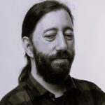 Imagem. Início da descrição de imagem: Retrato em tons de sépia ou preto e branco de um homem de pele clara, com barba cheia escura e cabelo longo escuro preso em rabo de cavalo na nuca. O homem está vestido com uma camisa xadrez escura e olha para baixo e para a direita, com uma expressão calma ou pensativa. O fundo é liso e claro, sem detalhes. Fim da descrição de imagem.