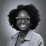 Imagem. Início da descrição de imagem: Retrato frontal em preto e branco de uma mulher negra sorridente com cabelos volumosos em estilo black power. Ela olha diretamente para a câmera, usa óculos de grau com armação escura e detalhes claros na parte superior, além de brincos de argola pequenos e um colar de corrente. Veste uma jaqueta de gola aberta sobre uma blusa de tom médio. O fundo é um gradiente cinza com iluminação centralizada atrás da cabeça, criando um efeito de auréola suave. Fim da descrição de imagem.