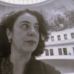 Descrição de imagem: um autorretrato em close-up e em tom sépia/preto e branco de uma mulher branca. Ela está olhando para o canto superior direito. Ela tem cabelos escuros e encaracolados e usa um brinco pequeno pendurado em formato geométrico. Veste uma camisa com gola desabotoada. O fundo é dramático, mostrando o teto abobadado de um grande salão ou museu, com um mural pintado (afresco) no domo e a arquitetura interna em estilo clássico em um plano inferior. Fim da descrição.