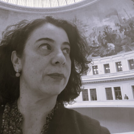 Descrição de imagem: um autorretrato em close-up e em tom sépia/preto e branco de uma mulher branca. Ela está olhando para o canto superior direito. Ela tem cabelos escuros e encaracolados e usa um brinco pequeno pendurado em formato geométrico. Veste uma camisa com gola desabotoada. O fundo é dramático, mostrando o teto abobadado de um grande salão ou museu, com um mural pintado (afresco) no domo e a arquitetura interna em estilo clássico em um plano inferior. Fim da descrição.