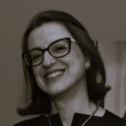 Descrição de imagem: um retrato em close-up e em tom sépia/preto e branco de uma mulher branca sorrindo amplamente. Ela tem cabelo liso e escuro na altura dos ombros e usa óculos de aro preto com formato cat-eye. Veste uma roupa escura (preta) e usa um colar delicado. O fundo é escuro e indefinido, com uma leve indicação de iluminação ambiente. Fim da descrição.