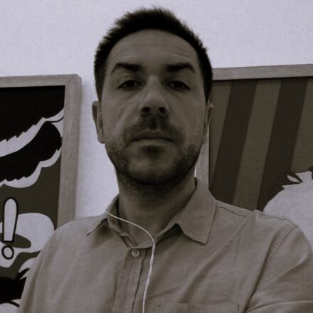 Descrição de imagem: um autorretrato em plano médio e em tom sépia/preto e branco de um homem branco olhando diretamente para a câmera com uma expressão neutra. Ele tem cabelo escuro curto e barba por fazer (barba stubble). Veste uma camisa de botão de cor clara e tem um fone de ouvido branco pendurado no pescoço. O fundo mostra uma parede clara e, de cada lado da cabeça, há o canto de uma obra de arte emoldurada com moldura de madeira; a da direita parece ter listras coloridas. Fim da descrição.