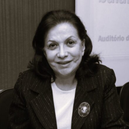 Descrição da imagem: Um retrato em plano médio e em tom sépia/preto e branco de uma mulher branca sentada e sorrindo. Ela tem cabelo escuro na altura dos ombros. Veste um blazer escuro sobre uma blusa branca. Um broche grande, que se assemelha a um camafeu com uma figura em relevo, está fixado na lapela do blazer. O fundo é composto por um banner ou parede com texto em português, incluindo a palavra "Auditório", indicando um evento ou conferência. Fim da descrição.