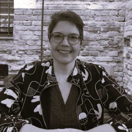 Descrição de imagem: um retrato em plano médio e em tom sépia/preto e branco de uma mulher branca sentada e sorrindo. Ela usa óculos de aro escuro e tem cabelo curto, um pouco acima das orelhas. Veste uma jaqueta escura com estampa abstrata em branco sobre uma blusa escura. Os braços estão dobrados sobre a superfície de uma mesa, na qual ela está apoiada. O fundo é uma parede de tijolos aparente. Fim da descrição.