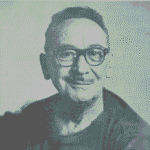 A imagem é um retrato em preto e branco de um homem de meia-idade, de pele clara, olhando diretamente para a câmera com um sorriso. Ele tem cabelo escuro e ralo penteado para trás. Usa óculos de armação escura e retangular. O homem tem um bigode fino. Ele veste uma camiseta escura com gola redonda. O fundo é uma parede clara e lisa, com uma leve textura granulada visível em toda a imagem (característica da fotografia). A iluminação é suave e uniforme, destacando o rosto sorridente do homem.