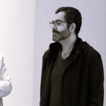 Imagem. Início da descrição de imagem: Retrato em plano médio, capturado em tom sépia, de um homem de pele clara, com barba e bigode escuros e cheios. Ele usa óculos de grau com armação fina e olha para a esquerda da câmera com uma expressão serena e contemplativa. Veste uma camiseta de gola em "V" escura sob uma jaqueta ou casaco aberto, também de cor escura e com textura sutil. À esquerda, vê-se a silhueta desfocada do braço de outra pessoa. O fundo é uma parede clara e lisa, característica de um ambiente de galeria ou museu. Fim da descrição de imagem.