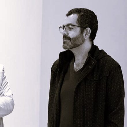 Imagem. Início da descrição de imagem: Retrato em plano médio, capturado em tom sépia, de um homem de pele clara, com barba e bigode escuros e cheios. Ele usa óculos de grau com armação fina e olha para a esquerda da câmera com uma expressão serena e contemplativa. Veste uma camiseta de gola em "V" escura sob uma jaqueta ou casaco aberto, também de cor escura e com textura sutil. À esquerda, vê-se a silhueta desfocada do braço de outra pessoa. O fundo é uma parede clara e lisa, característica de um ambiente de galeria ou museu. Fim da descrição de imagem.