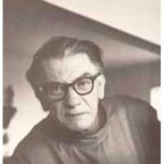 A imagem é um close-up em preto e branco de um homem de meia-idade ou idoso, de pele clara, olhando diretamente para a câmera com uma expressão neutra ou ligeiramente séria. Ele tem cabelo escuro e ralo, penteado para trás. Usa óculos de armação escura e arredondada. Há rugas visíveis na testa e ao redor dos olhos. O homem veste um suéter de gola rolê escuro. O fundo é uma parede clara e lisa, com uma parte escura visível no canto superior direito. A iluminação é suave, com a luz vindo da esquerda, criando um leve contraste de luz e sombra no rosto.