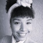 Imagem. Início da descrição de imagem: Retrato frontal em preto e branco de uma mulher negra sorridente com cabelos crespos. Ela usa uma faixa branca larga na cabeça, finalizada com um nó volumoso no topo, e possui um piercing de argola no septo. A mulher veste uma blusa branca de gola alta sob um casaco de tricô listrado em tons de cinza. Ela está com a mão direita tocando levemente o pescoço e olha diretamente para a câmera. O fundo é uma parede de textura granulada. Fim da descrição de imagem.