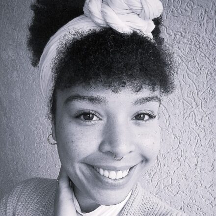 Imagem. Início da descrição de imagem: Retrato frontal em preto e branco de uma mulher negra sorridente com cabelos crespos. Ela usa uma faixa branca larga na cabeça, finalizada com um nó volumoso no topo, e possui um piercing de argola no septo. A mulher veste uma blusa branca de gola alta sob um casaco de tricô listrado em tons de cinza. Ela está com a mão direita tocando levemente o pescoço e olha diretamente para a câmera. O fundo é uma parede de textura granulada. Fim da descrição de imagem.