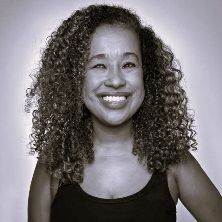 Descrição de imagem: A imagem é um retrato em preto e branco de uma mulher negra sorrindo amplamente, olhando ligeiramente para o lado esquerdo da imagem. Ela tem cabelos cacheados e volumosos, que emolduram seu rosto e se estendem para os lados. Seu sorriso é aberto, mostrando os dentes, e seus olhos estão levemente cerrados devido à intensidade da expressão. A mulher veste uma regata escura. O fundo da imagem é uma parede lisa e clara. A iluminação é brilhante, realçando o cabelo e a expressão feliz do rosto. Fim da descrição.