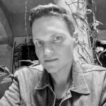 Descrição de imagem: Um retrato em close-up em preto e branco de um homem branco de olhos claros. Ele está olhando diretamente para a câmera. Ele tem cabelo curto, de tom médio e penteado. Sua expressão é séria e seus olhos estão fixos no observador. Ele veste uma jaqueta jeans com gola. O fundo é uma área externa sombreada com elementos arquitetônicos (uma coluna ou pilar) e plantas trepadeiras (hera ou similar) crescendo ao redor, criando um ambiente de pátio ou varanda. Fim da descrição.