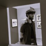 Descrição de imagem: Uma foto em preto e branco (sépia) de um homem branco de perfil. Ele está em pé dentro de uma galeria ou sala de exposição. A imagem é enquadrada por um portal ou abertura escura no primeiro plano. O homem tem cabelo liso na altura dos olhos e barba cheia, veste uma camisa de manga curta escura aberta sobre uma camiseta preta e um colar fino. Ele está olhando para a direita e tem as mãos nas costas. Ao fundo, nas paredes da galeria, há várias obras de arte emolduradas penduradas. Fim da descrição.