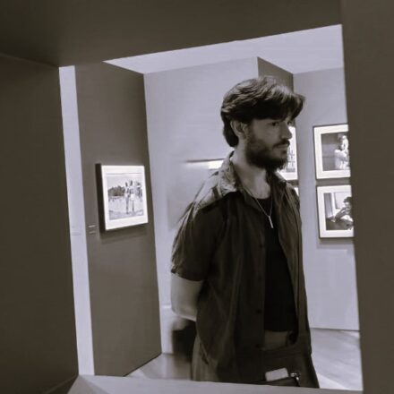 Descrição de imagem: Uma foto em preto e branco (sépia) de um homem branco de perfil. Ele está em pé dentro de uma galeria ou sala de exposição. A imagem é enquadrada por um portal ou abertura escura no primeiro plano. O homem tem cabelo liso na altura dos olhos e barba cheia, veste uma camisa de manga curta escura aberta sobre uma camiseta preta e um colar fino. Ele está olhando para a direita e tem as mãos nas costas. Ao fundo, nas paredes da galeria, há várias obras de arte emolduradas penduradas. Fim da descrição.