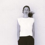 Descrição de imagem: um retrato em preto e branco (com leve tonalidade sépia) de uma mulher branca em plano médio. Ela está posicionada centralmente contra uma parede clara e texturizada que ocupa a maior parte do fundo. A mulher tem cabelos escuros, presos em um rabo de cavalo que está levemente esvoaçado à direita. Ela sorri sutilmente e usa um batom escuro. Veste uma camiseta branca de manga curta e ombros estruturados (estilo muscle tee) e calças escuras (pretas ou grafite), com as mãos nos bolsos. Fim da descrição.