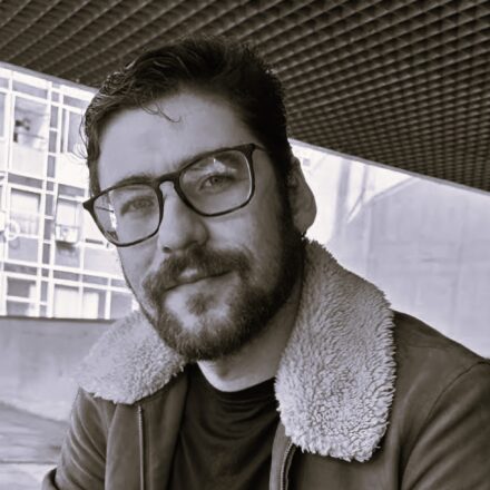 Descrição de imagem: Um retrato em close-up em preto e branco (com leve tonalidade sépia) de um homem branco olhando diretamente para a câmera com uma expressão séria. Ele tem barba e bigode cheios, e usa óculos de aro preto grosso. Ele veste uma jaqueta de camurça com um colarinho de pele de carneiro (sherpa) bege ou branco. O fundo sugere um ambiente urbano ou arquitetônico, com uma estrutura de teto vazado (cobogó ou brise) escuro na parte superior e um prédio moderno de janelas quadradas em segundo plano, parcialmente desfocado. Fim da descrição.