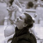 Imagem. Início da descrição de imagem: Retrato de perfil em tom sépia de uma mulher sorridente com o rosto voltado para cima. Ela tem a pele clara, olhos semicerrados pelo sorriso e cabelos escuros presos em um coque alto. Veste um casaco escuro acolchoado e um cachecol volumoso da mesma cor. O fundo da imagem está desfocado, mostrando o que parece ser uma escultura clássica em um ambiente interno. Fim da descrição de imagem.