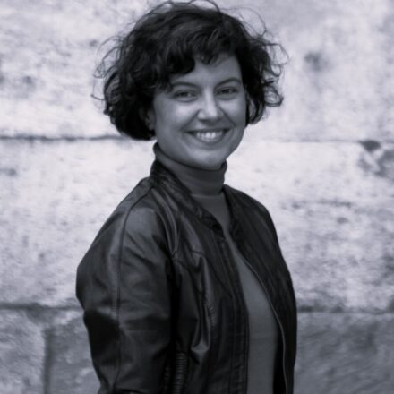 Imagem. Descrição da imagem: Um retrato fotográfico em preto e branco focado no plano médio mostra uma mulher de pele clara sorrindo. Ela está ligeiramente de perfil esquerdo, com o rosto voltado diretamente para a câmera. Seu cabelo é castanho-escuro, curto, volumoso, com cachos abertos e uma franja sutil sobre a testa. Ela exibe um sorriso largo que revela os dentes superiores. A mulher veste uma blusa de gola alta escura sob uma jaqueta de material sintético semelhante a couro, aberta na frente, com costuras marcadas e acabamentos canelados na gola e na barra. O fundo é composto por uma parede rústica de blocos ou pedras claras, com texturas ásperas e imperfeições naturais que estão ligeiramente fora de foco. A iluminação é difusa e uniforme, destacando os contornos de seu rosto e o brilho suave da jaqueta. Fim da descrição.