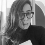 Imagem. Início da descrição de imagem: Retrato em close-up em preto e branco de uma mulher de cabelos longos e escuros, que cobrem parcialmente um lado do seu rosto. Ela usa óculos de armação grossa e escura de formato angular (estilo gatinho ou máscara) e veste uma blusa preta de gola alta. Ela mantém uma expressão séria e o fundo da imagem é desfocado e escuro. Fim da descrição de imagem.