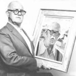 A imagem é uma fotografia em preto e branco de um homem idoso que segura um autorretrato emoldurado. O homem está voltado para o espectador. Ele é careca e usa óculos escuros de armação grossa e preta; veste um terno escuro e uma camisa clara por baixo. Sua expressão é séria. Com as duas mãos, ele segura um autorretrato emoldurado. O fundo da imagem é uma parede clara e lisa, que ajuda a destacar o homem e o quadro. Há um contraste forte entre os tons escuros da roupa do homem e os óculos, e os tons mais claros do fundo e da camisa. A imagem cria um efeito de "quadro dentro do quadro", com o sujeito segurando uma representação de si mesmo ou de uma figura muito semelhante