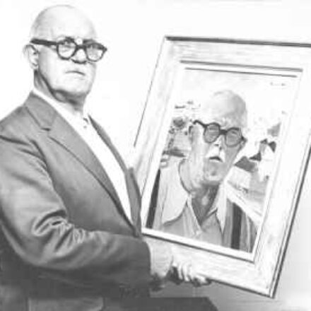 A imagem é uma fotografia em preto e branco de um homem idoso que segura um autorretrato emoldurado. O homem está voltado para o espectador. Ele é careca e usa óculos escuros de armação grossa e preta; veste um terno escuro e uma camisa clara por baixo. Sua expressão é séria. Com as duas mãos, ele segura um autorretrato emoldurado. O fundo da imagem é uma parede clara e lisa, que ajuda a destacar o homem e o quadro. Há um contraste forte entre os tons escuros da roupa do homem e os óculos, e os tons mais claros do fundo e da camisa. A imagem cria um efeito de "quadro dentro do quadro", com o sujeito segurando uma representação de si mesmo ou de uma figura muito semelhante