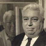 A imagem é um retrato em preto e branco de um homem de meia-idade, com cabelos brancos e curtos. Ele está vestido com um terno escuro e uma gravata preta. O homem tem um rosto arredondado e uma expressão neutra, olhando diretamente para a câmera. Atrás dele, ligeiramente à esquerda e desfocado, há um quadro emoldurado com um retrato. O retrato dentro do quadro mostra um homem com características muito semelhantes ao sujeito em primeiro plano, incluindo cabelos brancos e rosto arredondado. O fundo do retrato parece ser uma parede de tom claro. A composição da imagem cria um efeito de "quadro dentro do quadro" ou de reflexão, com o homem em primeiro plano e sua possível representação artística ao fundo.