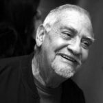 A imagem é um close-up em preto e branco de um homem idoso sorrindo. Ele tem cabelos curtos e grisalhos, com entradas visíveis, e uma barba e bigode também grisalhos. A boca está aberta em um sorriso, mostrando os dentes. Seus olhos estão ligeiramente semicerrados. O homem veste uma jaqueta escura e uma camiseta mais clara por baixo. O fundo é escuro e desfocado.