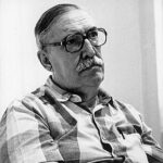 A imagem é um retrato em preto e branco de um homem de meia-idade ou idoso, que está com a cabeça inclinada para a esquerda e olha ligeiramente para sua direita, com uma expressão séria. O homem, de pele clara, possui um tipo corporal robusto e cabelo escuro e curto nas laterais. Tem um bigode proeminente e usa óculos de armação escura e quadrada. Ele veste uma camisa de manga curta com um padrão xadrez claro e escuro. Seus braços estão cruzados na frente do corpo. Um objeto fino, semelhante a uma caneta, está preso no bolso da camisa. O fundo é branco e liso, sem detalhes perceptíveis.