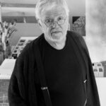 A imagem é um retrato em preto e branco de um homem idoso, posicionado da cintura para cima. Ele possui cabelos brancos, barba branca e densa, e usa óculos de armação fina e redonda. Seus olhos são escuros e direcionados para a câmera. O homem veste uma camiseta de gola redonda escura, um cardigã escuro aberto sobre a camiseta, calças escuras, um cinto escuro com fivela retangular e suspensórios pretos com prendedores metálicos prateados. Sua mão esquerda está no bolso da calça. O fundo da imagem apresenta elementos de um ambiente interno. À esquerda, são visíveis recipientes com pincéis e materiais, além de uma estrutura com múltiplas divisórias. À direita, uma superfície com um padrão texturizado e linear é parcialmente visível. A iluminação é difusa, realçando os contrastes de preto, branco e tons de cinza.