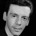 A imagem é um close-up em preto e branco de um homem jovem, de pele clara, olhando diretamente para a câmera com uma expressão neutra ou ligeiramente sorridente. Ele tem cabelo escuro, curto e penteado para trás. O homem veste uma camisa escura, com a gola visível. O fundo é completamente escuro e liso, sem detalhes discerníveis, o que destaca o rosto do homem. A iluminação é frontal e suave, criando alguns reflexos nos cabelos e no rosto.