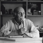 A imagem é um retrato em preto e branco de um homem idoso, sentado em uma mesa e olhando para sua direita, fora do enquadramento. O homem, de pele clara, é calvo na parte superior da cabeça, com cabelo grisalho e esparso nas laterais. Ele tem um bigode e sobrancelhas proeminentes. Sua expressão é pensativa. Ele veste uma camisa de manga comprida e clara, com as mangas dobradas na altura dos antebraços. Seus braços estão apoiados na mesa, e ele segura um objeto fino e escuro (possivelmente uma caneta ou lápis) entre os dedos e a boca, tocando o queixo ou lábios. Sobre a mesa branca em primeiro plano, há um objeto longo e cilíndrico, que parece ser um rolo de papel ou um tubo de desenho, e uma régua ou instrumento de medição. O fundo da imagem é uma estante cheia de objetos diversos, incluindo caixas, potes e livros, em prateleiras escuras. A iluminação vem da esquerda, criando sombras que acentuam as rugas do rosto e a profundidade do ambiente.