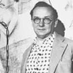 A imagem é um retrato em preto e branco de um homem de meia-idade, posicionado da cintura para cima, olhando diretamente para a câmera. Ele tem cabelo escuro, penteado para trás, e usa óculos redondos de armação escura. O homem veste uma camisa clara com um padrão de bolinhas escuras e um paletó de cor clara por cima. O fundo da imagem, parcialmente visível à esquerda, parece ser uma parede com uma obra de arte ou um desenho em estilo figurativo, com linhas escuras e formas abstratas ou humanas. A iluminação é uniforme, destacando o rosto e a textura das roupas.