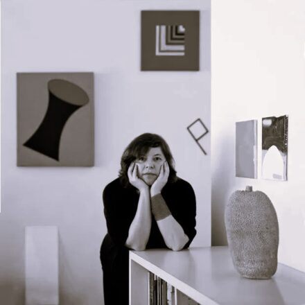 Imagem. Início da descrição de imagem: Retrato frontal em tom sépia de uma mulher de pele clara e cabelos escuros curtos, na altura do queixo. Ela está posicionada no centro, apoiando o queixo nas palmas das mãos, com os cotovelos sobre um móvel branco. Usa uma blusa escura e uma pulseira larga no pulso esquerdo. Ao fundo, em uma parede clara, veem-se diversas obras de arte geométricas emolduradas de diferentes tamanhos e formatos. Sobre o móvel à direita, em primeiro plano, destaca-se uma escultura que mimetiza a forma de uma jaca, característica de suas investigações sobre objetos cotidianos. A iluminação é suave, criando uma atmosfera introspectiva e artística. Fim da descrição de imagem.