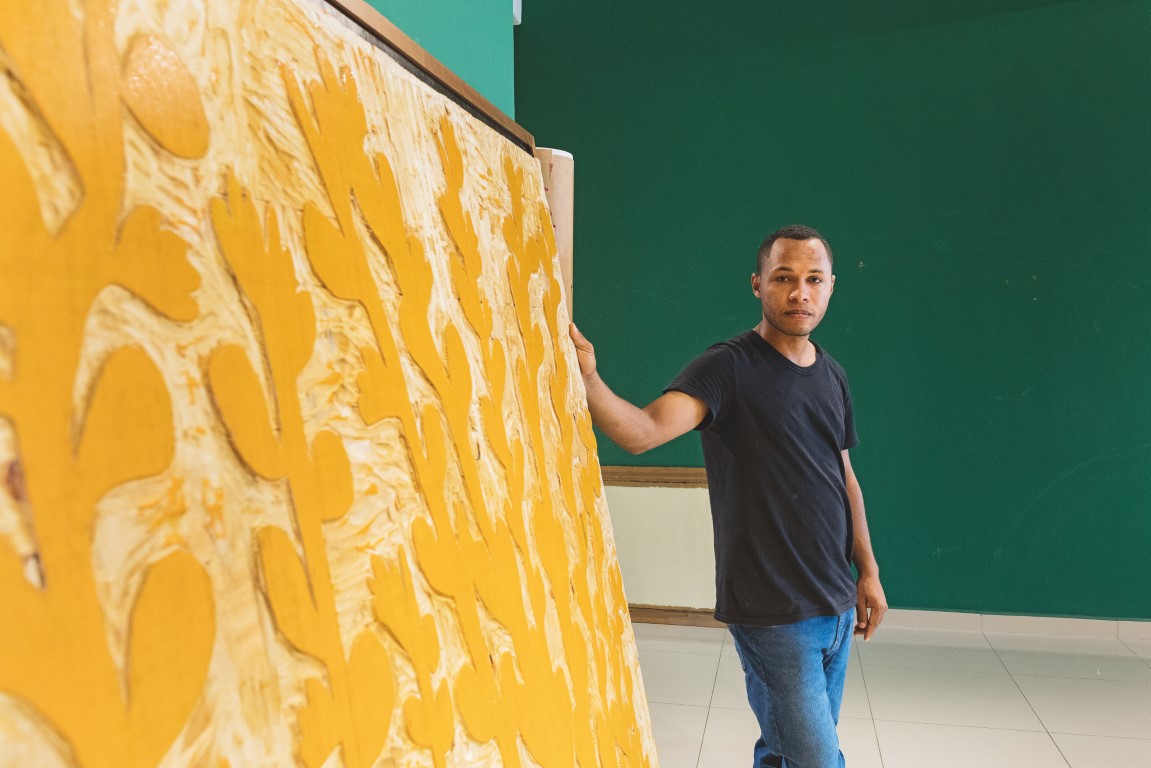 Descrição da imagem: fotografia horizontal de Santídio Pereira em seu ateliê. Ele está em pé, de frente para a esquerda, segurando com a mão direita uma matriz de xilogravura. O ateliê tem chão beje e parede verde. A matriz retangular, é da altura dele, com quase 1,70m de altura. É tingida de amarelo, com desenhos de caules e folhas gravados. Fim da descrição da imagem.