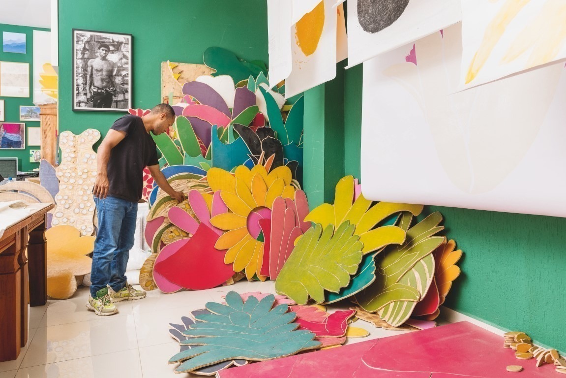 Descrição da imagem: fotografia horizontal de Santídio Pereira trabalhando em seu ateliê. Ele está em pé, de frente para a direita, observando diversas matrizes de xilogravuras coloridas apoiadas umas sobre as outras. As peças de xilogravura são grandes e em formatos de folhas e flores. Há trabalhos impressos pendurados no teto, no lado direito da imagem, e uma fotografia pendurada na parede ao fundo. Fim da descrição da imagem.