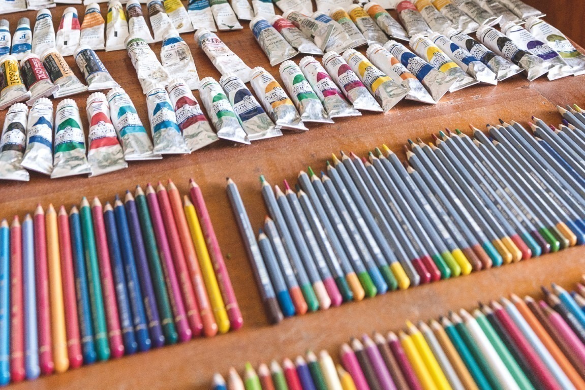 Descrição da imagem: fotografia horizontal que exibe uma mesa de madeira com diversas bisnagas de tintas e lápis coloridos sobre ela. Há mais de 50 tubos e lápis, dispostos em fileiras e organizados por cor. Há lápis e tubos amarelos, vermelhos, verdes, azuis, de diversos tons, entre outras cores. Fim da descrição da imagem.