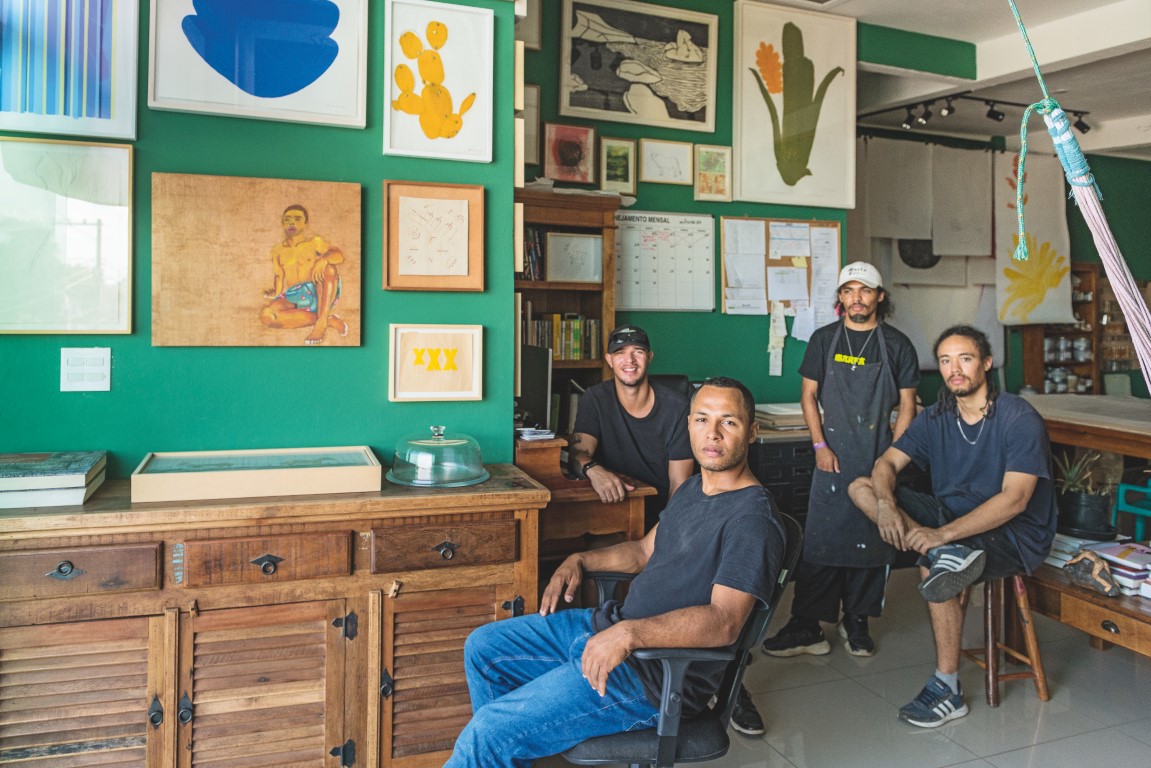 Descrição da imagem: fotografia horizontal de Santídio Pereira no ateliê, sentado com os braços apoiados no encosto da cadeira à frente de três homens. Santídio usa camiseta preta e calça jeans azul. Atrás dele, da esquerda para direita: um homem branco sentado. Usa boné preto e camiseta preta; um homem negro de pé. Usa boné branco, camisa e avental pretos; e por fim, um homem negro sentado. Usa os cabelos trançados, camisa azul escuro e bermuda preta. Nas paredes, há diversos quadros e desenhos pendurados. Há armários de canto, mesas e bancadas ao longo do ateliê. No canto superior direito da imagem, é possível perceber a ponta de uma rede de dormir armada, pendendo do teto. Fim da descrição da imagem.