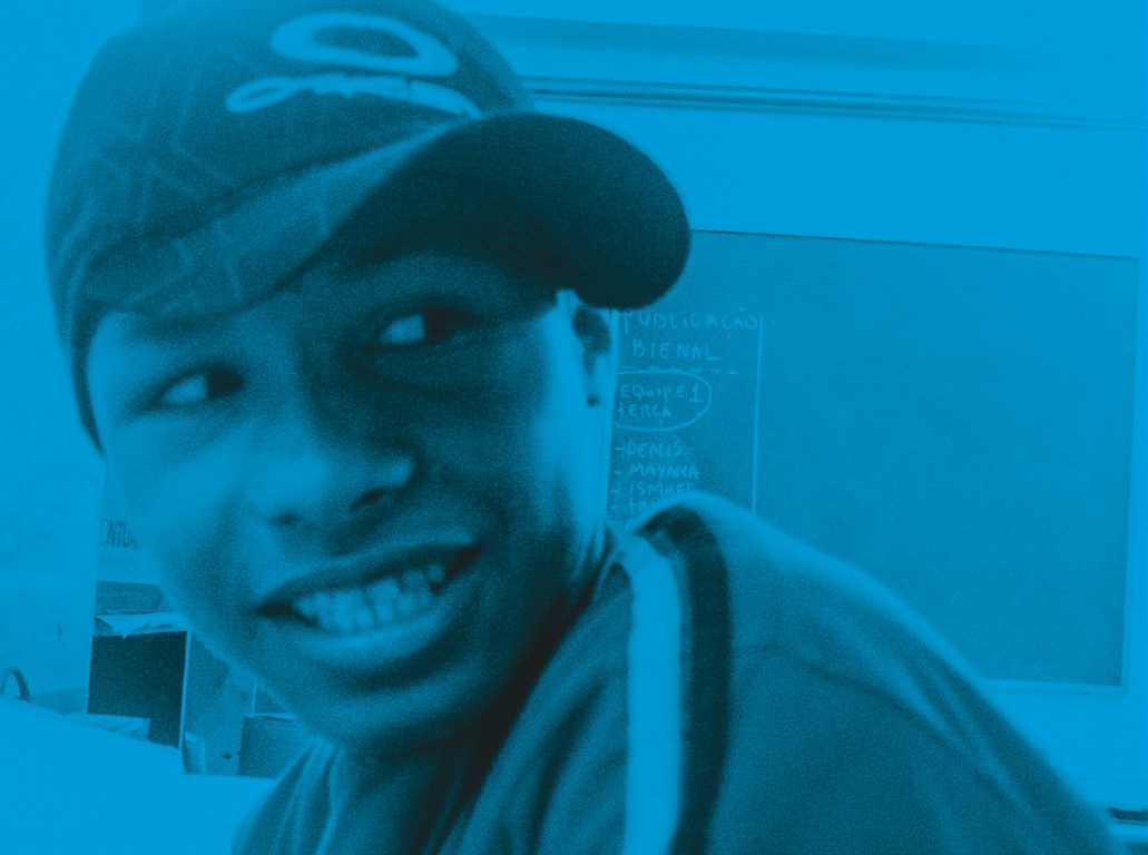 Descrição da imagem: fotografia horizontal com filtro azul e efeito monocromático. O retrato, um pouco desfocado, é de Santídio mais jovem, sentado em uma sala de aula. Sorridente, ele olha para a esquerda, Usa boné com a aba curva e virado para a direita e camisa com um par de listras nos ombros. Fim da descrição da imagem.