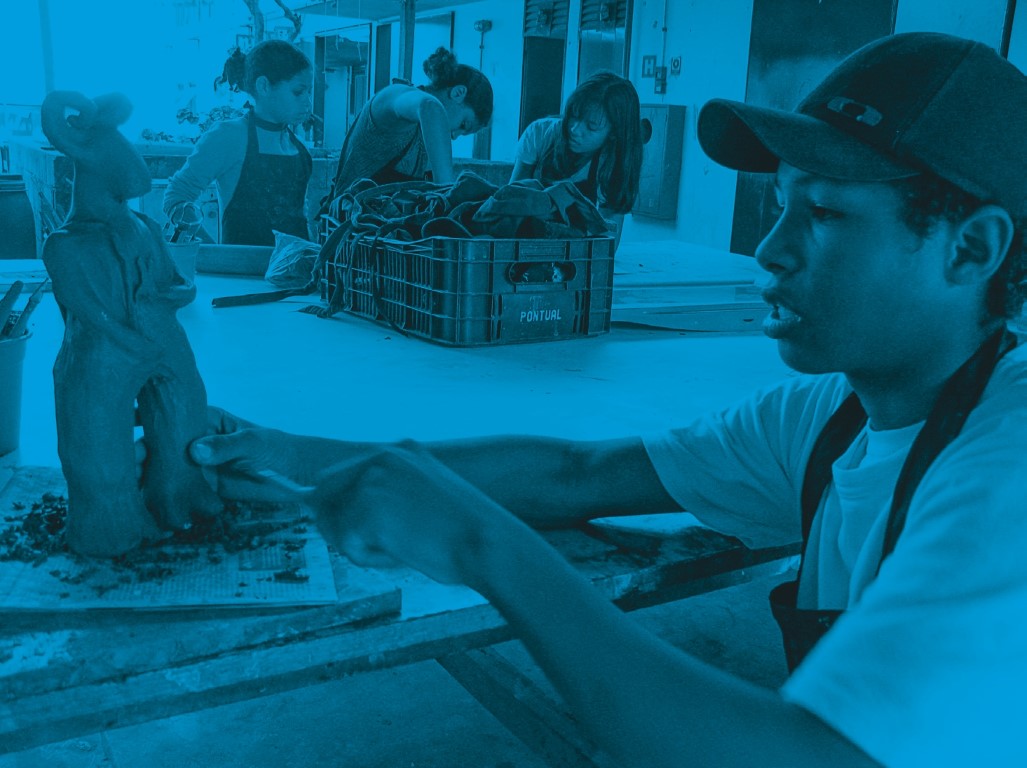 Descrição da imagem: fotografia colorida horizontal com filtro azul. Santídio está sentado a uma mesa em um ateliê de esculturas. Usa boné e avental pretos. Ele modela uma peça de argila, que é um híbrido de bode e humanoide. Ao redor da mesa, ao fundo, três garotas de pé e também de avental, modelam outra peça. Uma caixa de feira sobre a mesa tem outros aventais dentro dela. Fim da descrição da imagem.