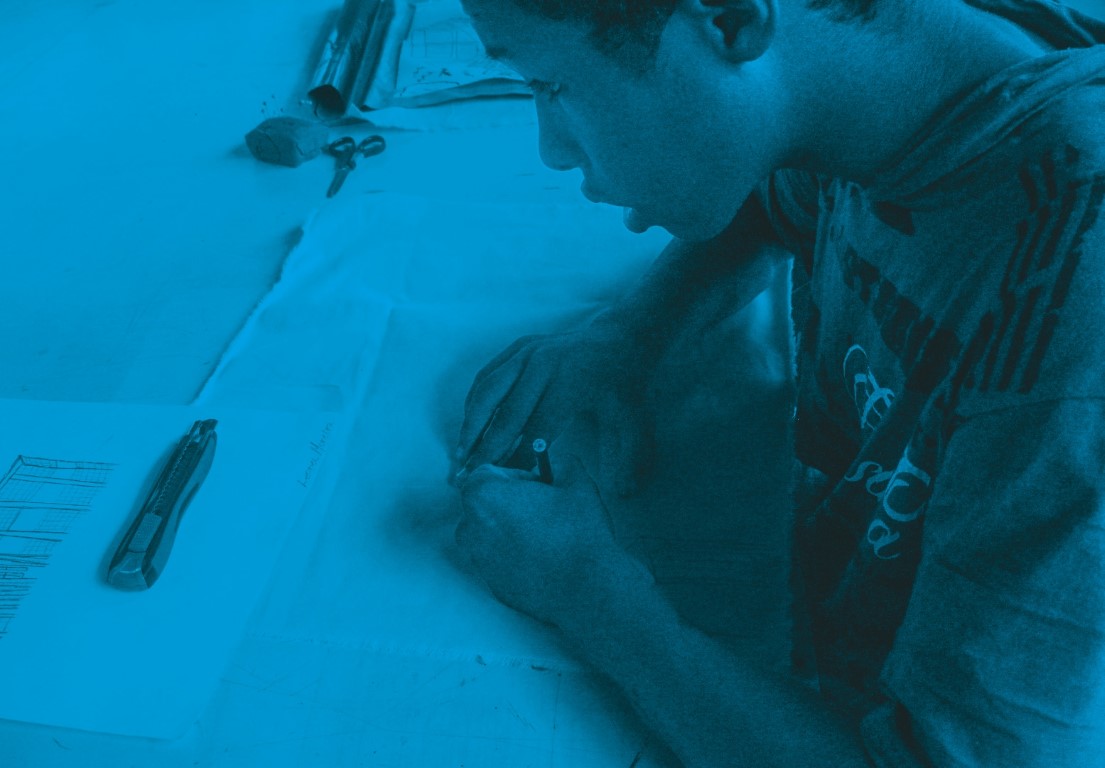 Descrição da imagem: fotografia colorida horizontal com filtro azul. Santídio desenha sobre um papel. Ao fundo, há outros papéis, um estilete, uma tesoura, um pedaço de argila e outros objetos sobre a mesa. Fim da descrição da imagem.