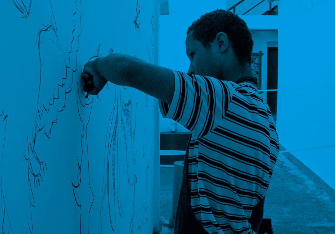Descrição da imagem: fotografia colorida horizontal com filtro azul. Santídio está de pé, de perfil para a esquerda, pinta um mural em uma parede branca com um giz preto ou carvão. Ele veste uma camisa listrada e avental preto. Fim da descrição da imagem.