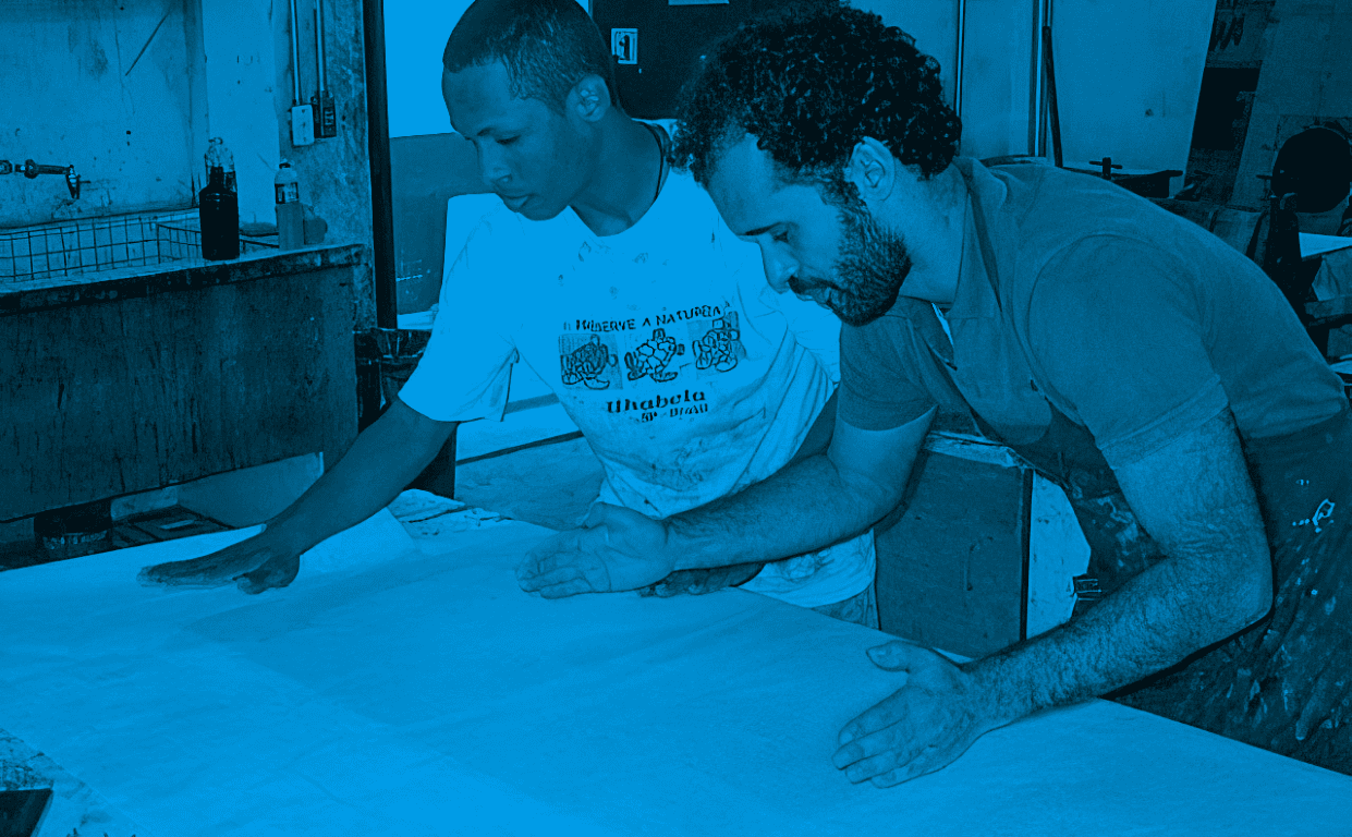 Descrição da imagem: fotografia colorida horizontal com filtro azul. Santídio e seu professor Fabrício estão de frente para uma mesa, com as mãos espalmadas sobre uma folha branca. Santídio usa camiseta branca com estampas de tartaruga. Ao lado, Fabrício usa barba, cabelo cacheado e camiseta de manga curta. Eles estão em um ateliê. Fim da descrição da imagem.