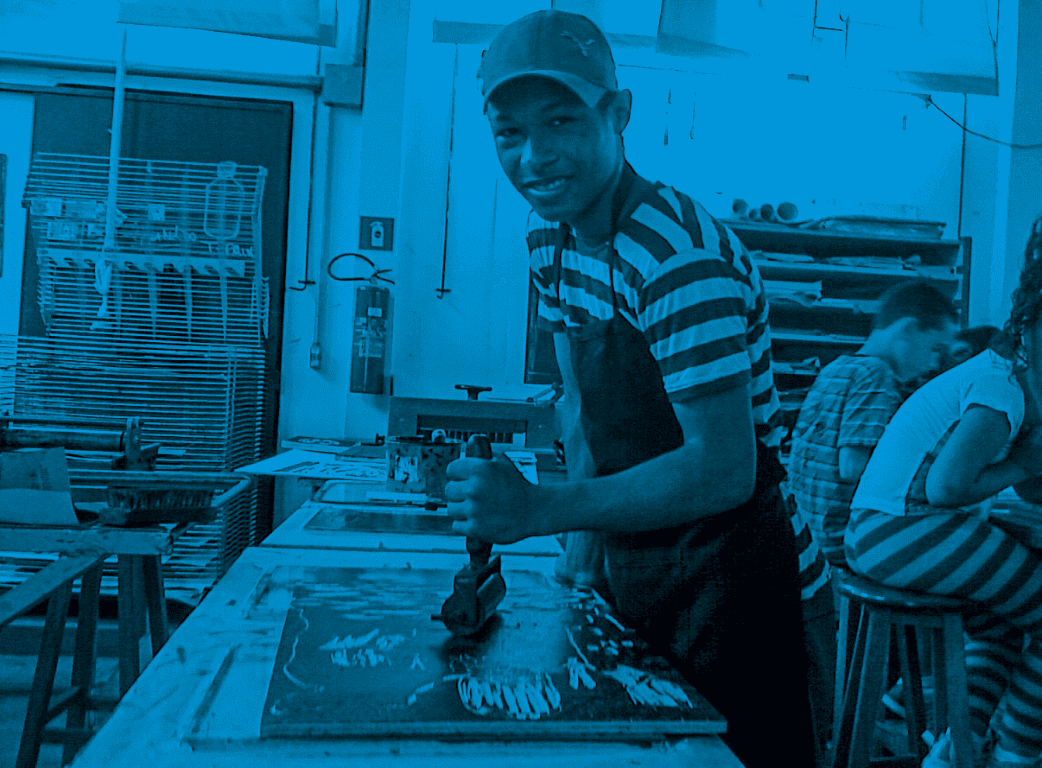Descrição da imagem: fotografia horizontal colorida com filtro azul. Santídio está em um ateliê de gravura. Ele usa boné e avental pretos, e está em pé, sorrindo, em frente a uma mesa com uma matriz de xilogravura, na qual trabalha usando um rolo de borracha. Há outras pessoas ao fundo trabalhando em outras mesas. Fim da descrição da imagem.