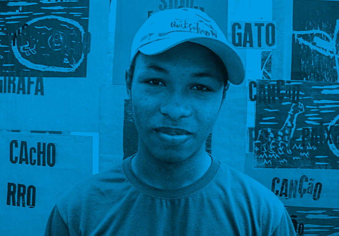 Descrição da imagem: fotografia horizontal colorida com filtro azul. Retrato de Santídio. Ele está em frente a uma parede com lambe-lambes de desenhos em xilogravura. Nelas, lê-se as palavras como "gato", "canção", "cacho", entre outras. Ele usa boné e camiseta em tons claros. Fim da descrição da imagem.